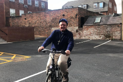 Revd Richard Teal smiles whilst riding his bicycle, surrounded by redbrick buildings. 