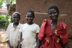 Beth stands to the right of her son and daughter, outside her home in Uganda 
