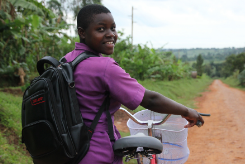 A young girl in a purple school uniform smiles with her bicycle, ready to travel to school. 