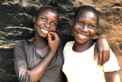 Two twin sisters embrace, standing outside their home in Uganda.