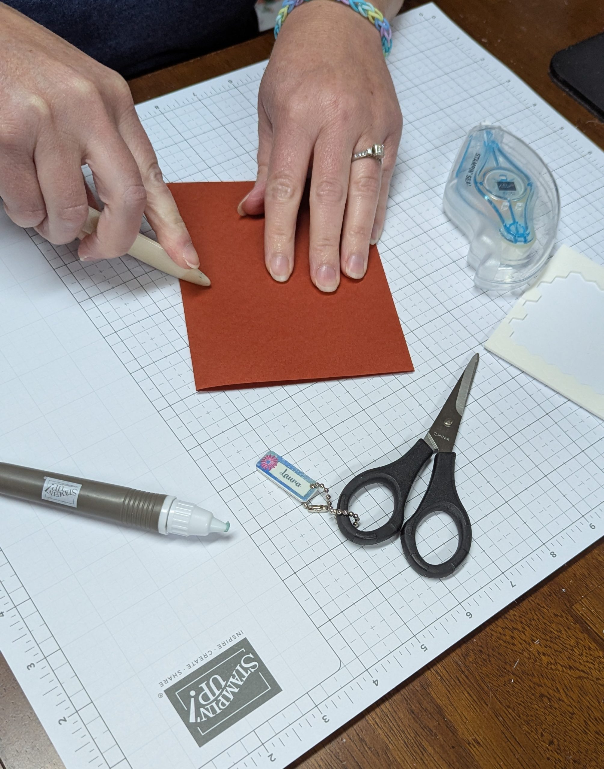 Hands scoring and folding dusty rose cardstock with a bone folder beside a half-finished handmade card