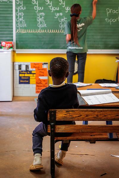 Young learner sitting at desk while teacher writes on blackboard