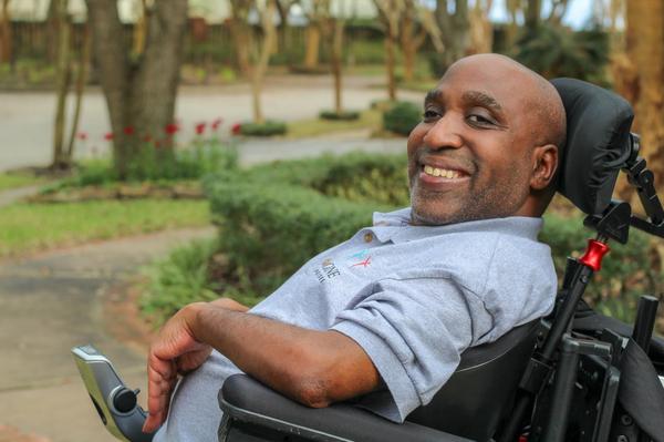 An image of Ricky Broussard, who is smiling and seated in his power wheelchair. He is wearing a gray polo
shirt with the Imagine Enterprises logo on it. In the background, there are out-of-focus tree trunks and a sidewalk lined with green shrubs. One of the trees has tall red flowers surrounding its base. 