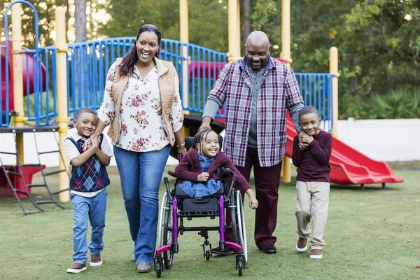 A happy African-American family with three young children walking together on a playground. The daughter, 5 years old, is in a wheelchair. She has
caudal regression syndrome, a rare congenital disorder which affects the development of the lower spine. She is in the middle between the parents. Her brothers, twin 4 year old boys, are on the ends, holding their parents' hands.