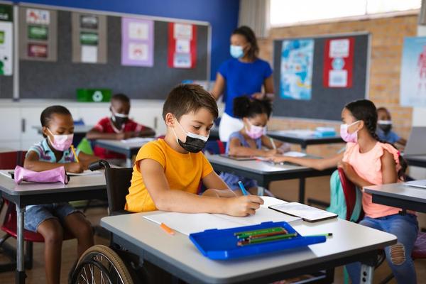 Elementary school students are seated at their desks. The main subject is is a young boy in his wheelchair seated at his desk, wearing a black mask and writing on a blank piece of paper with a blue marker.