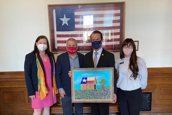 The Arc of Texas CEO Jennifer Martinez, The Arc of Texas President Gary Hidalgo, Speaker of the Texas House Dade Phelan, and The Arc of Greater
Beaumont CEO Sarah Hardin are standing in a row under a framed Texas naval flag. Everyone is wearing a face mask. Speaker Phelan is holding a painting by Ashley Sattler. The painting is of a brown barn with a Texas flag painted on the roof. The sky is bright blue and the ground is covered with colorful wildflowers. There is a tree next to the barn. 