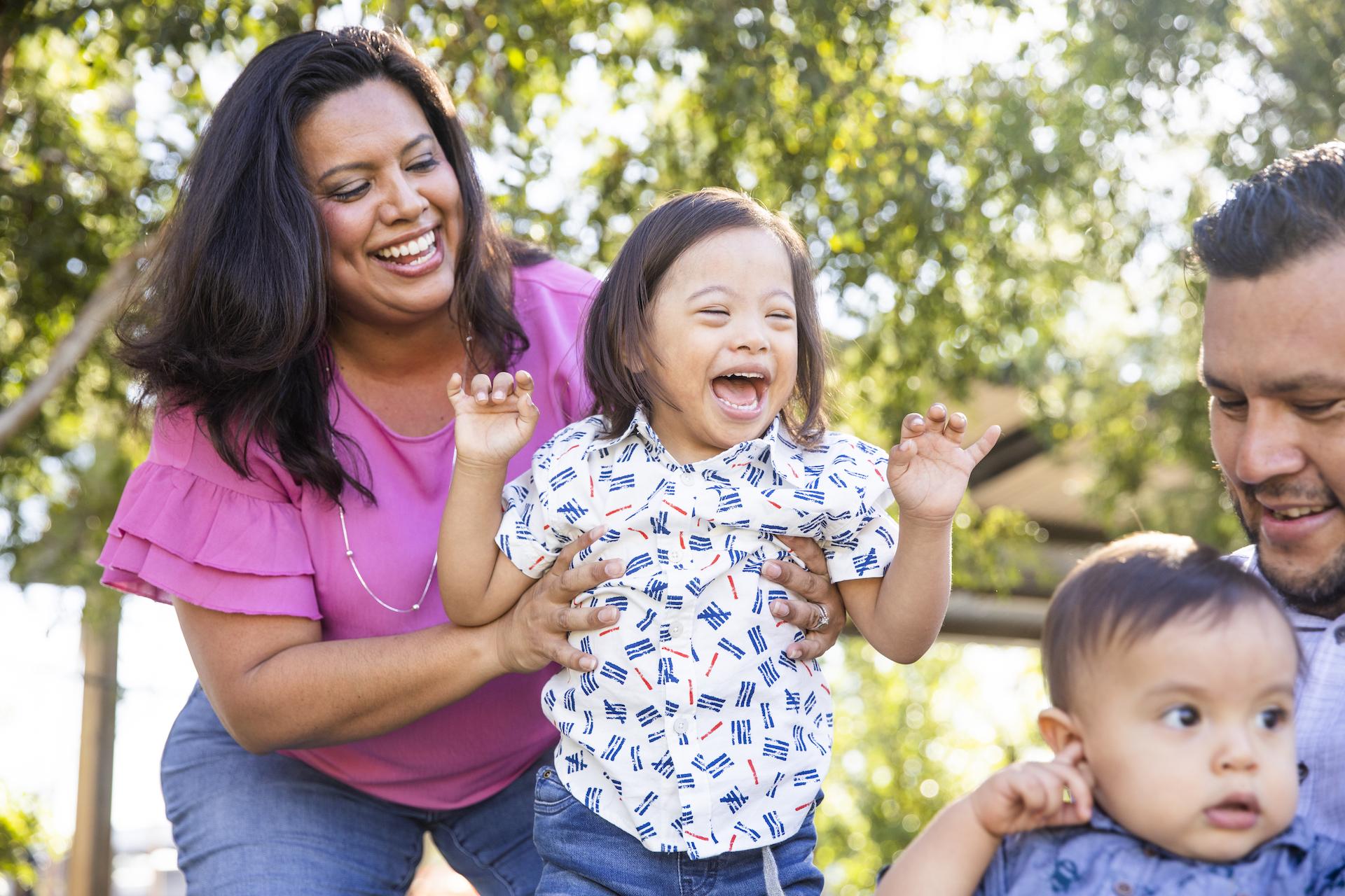 A mom holds her son. The young boy has down's syndrome. His younger brother is being held by his father. They are all playing outdoors and smiling.