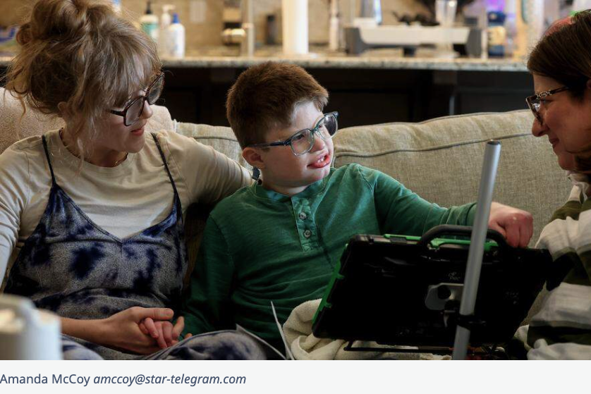 Luke sits with a smile on his face with his mother and caretaker on the couch at their home while using a communication tablet to interact.