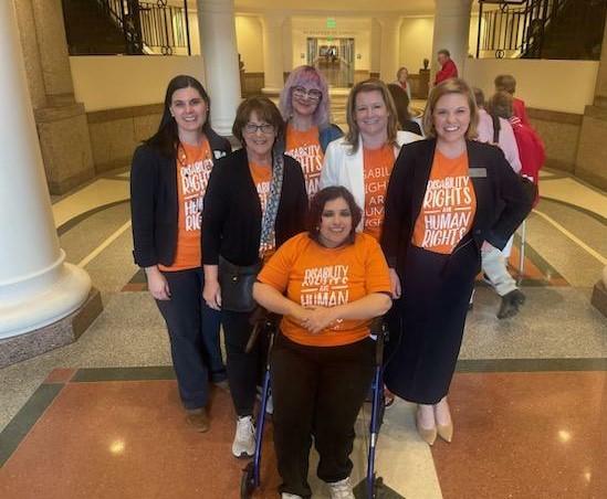 A picture of Ashley Ford with Veronica Ayala and other self-advocates at the Texas Capitol. They are wearing orange shirts with white text that say, "Disability rights are human rights."