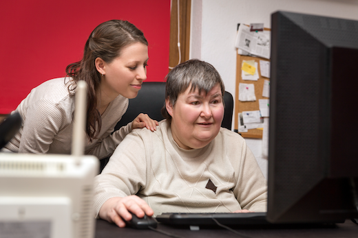 A woman with an intellectual disability is seated and looking at a computer with her hand on the mouse. Her direct care worker is standing behind her with her hand on the woman's shoulder. 