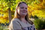 Photo of Shelbi Davenport smiling, sitting in a chair. Background is of fall trees.