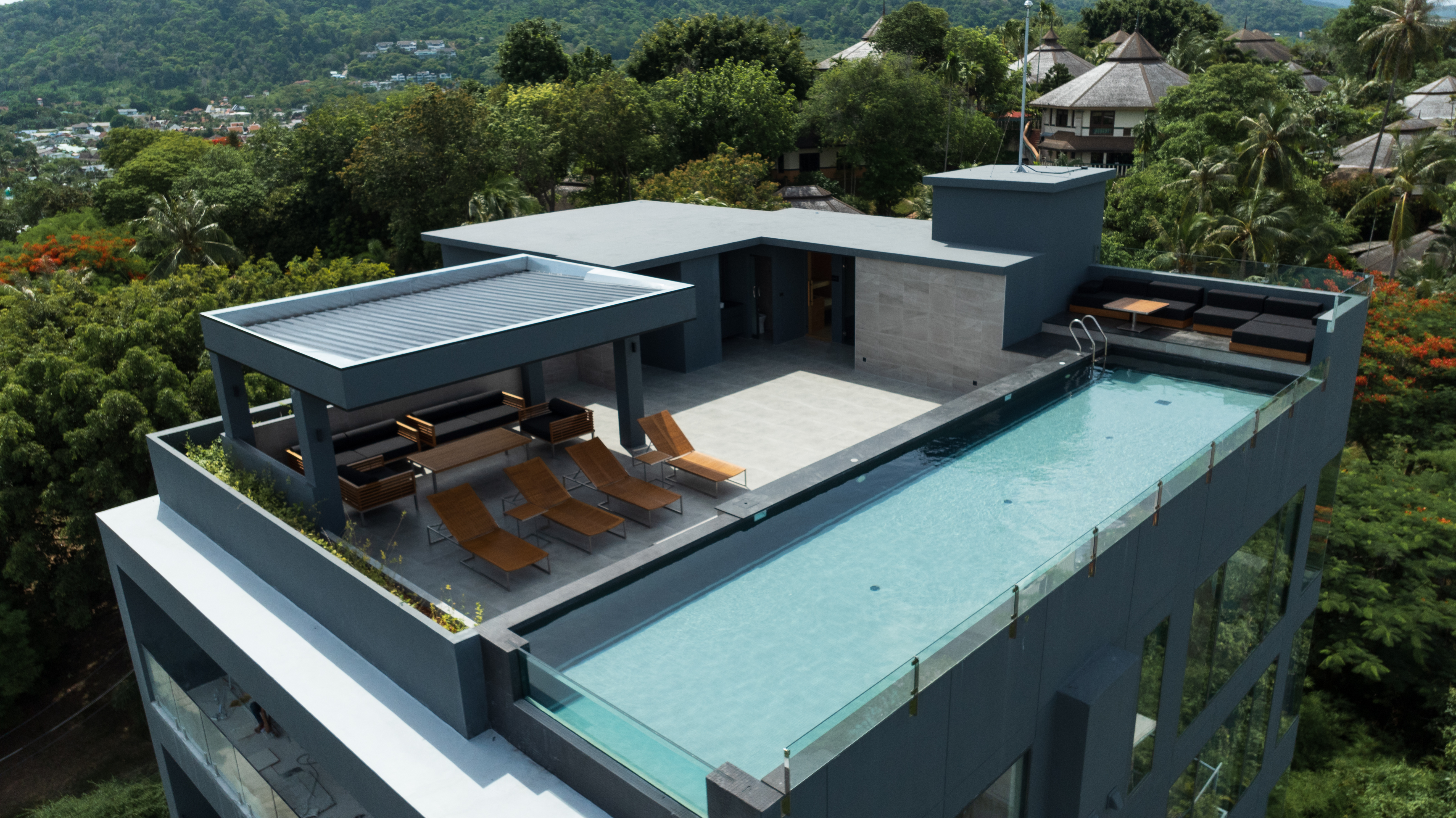 Aerial view of a rooftop infinity pool and lounge terrace surrounded by green hills in Phuket