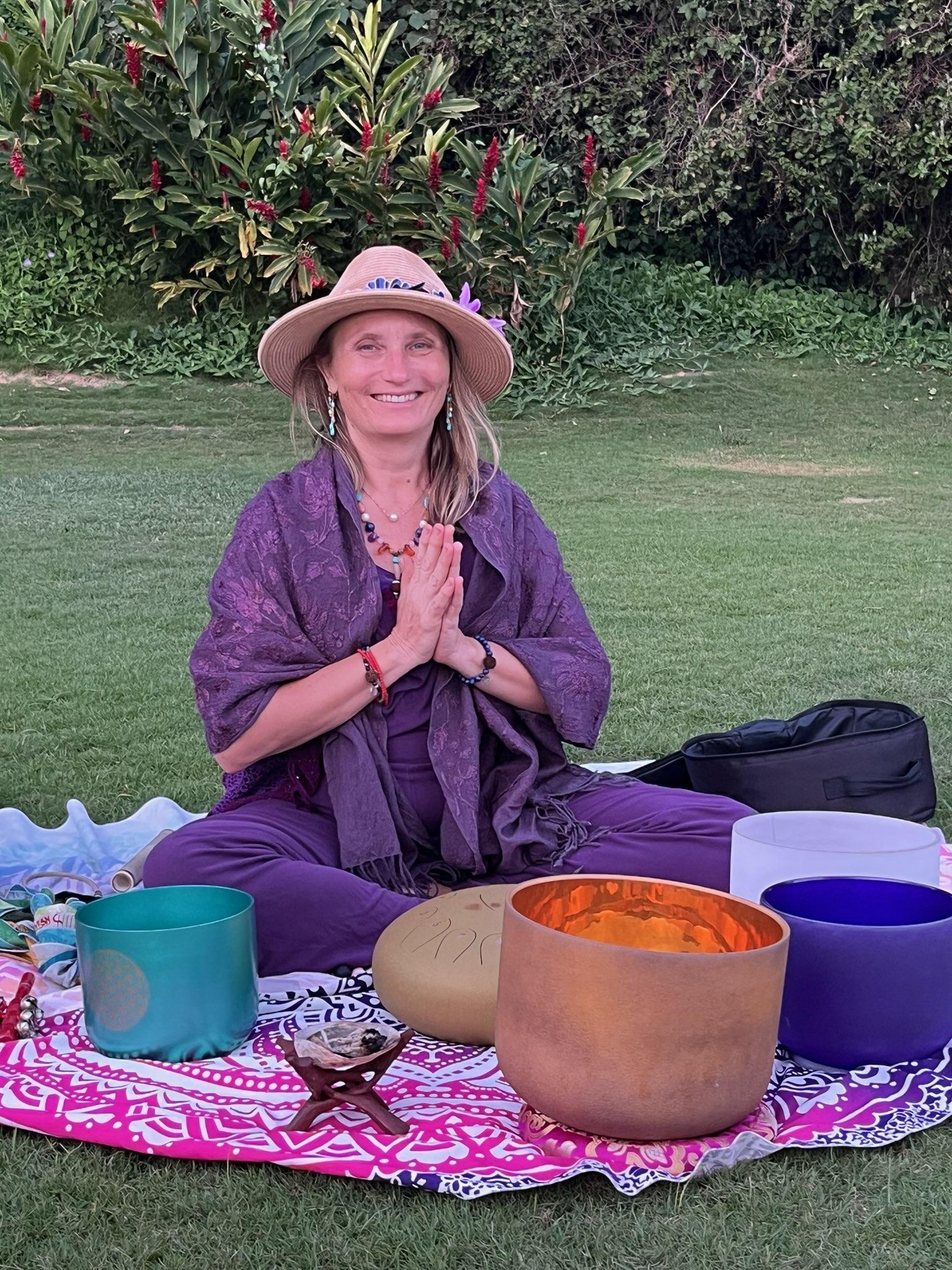 Anya Love smiling in hands-to-heart prayer pose on the grass, surrounded by her crystal singing bowls