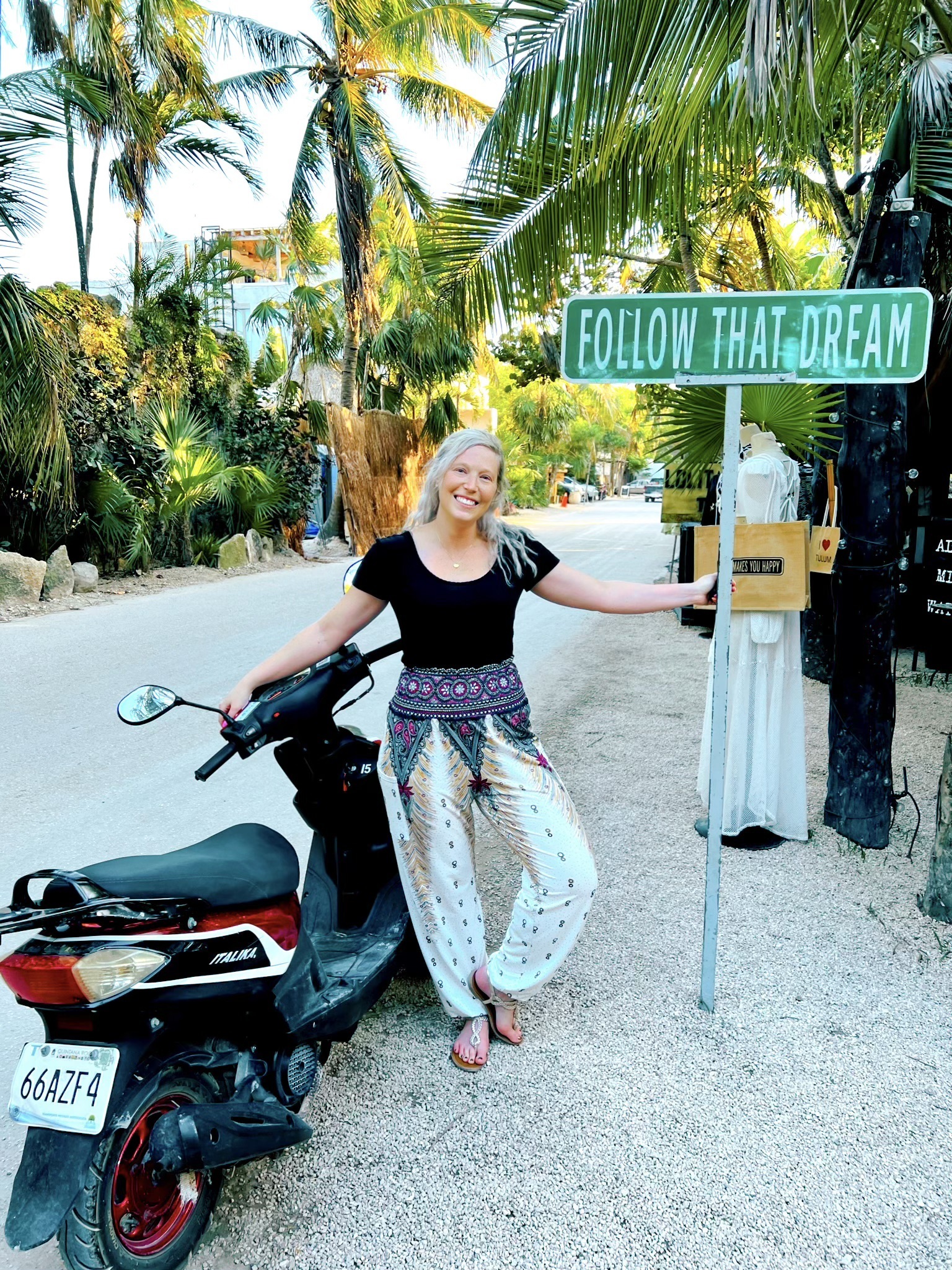 A smiling woman beside a scooter under palm trees and a FOLLOW THAT DREAM street sign in Tulum
