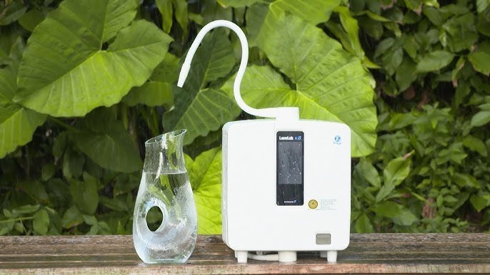 A Kangen water ionizer machine and a glass carafe of fresh water on a wooden table with greenery behind