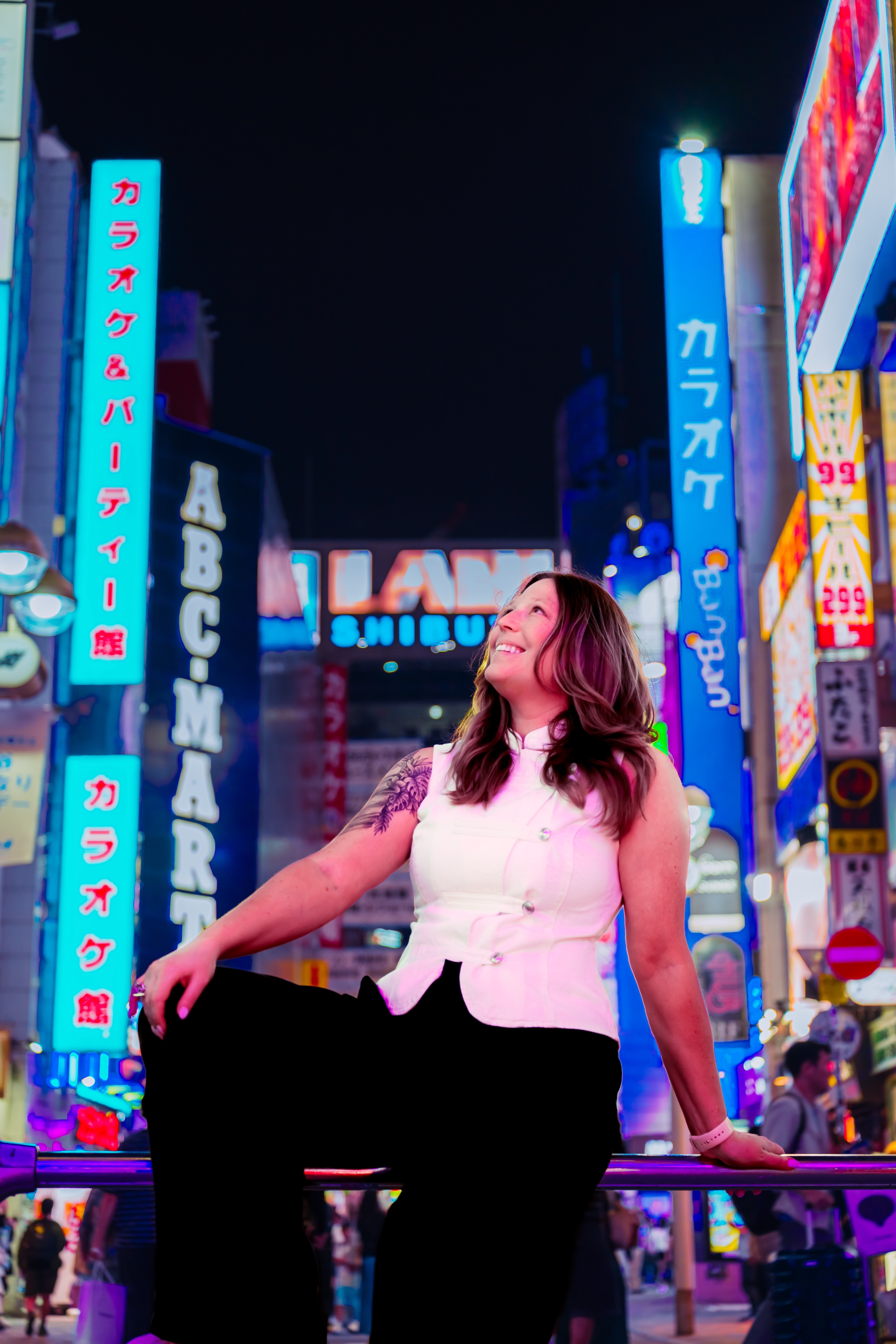 A woman sitting on a railing beneath glowing Shibuya signs, looking upward