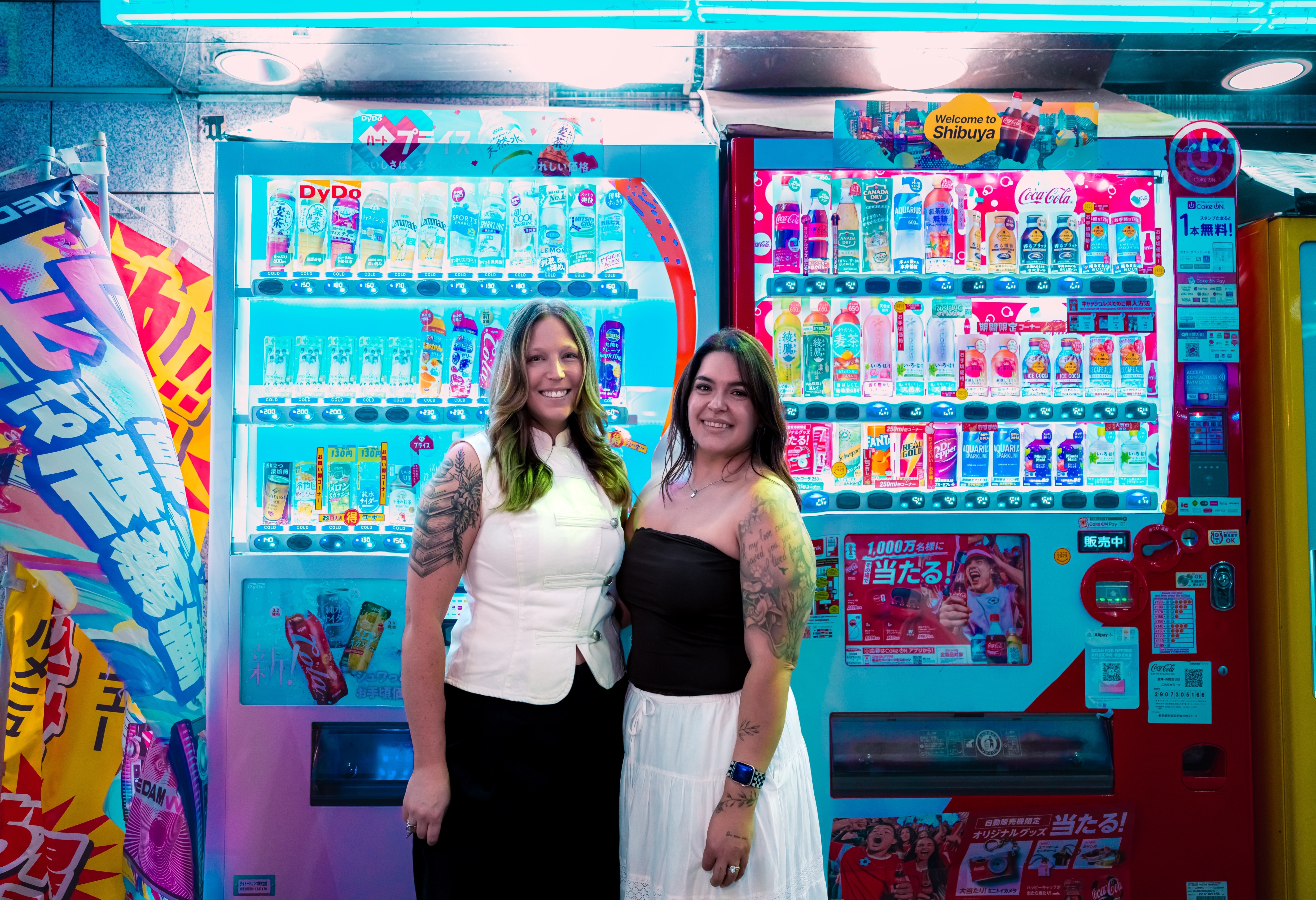 Two women smiling together in front of brightly lit Japanese vending machines