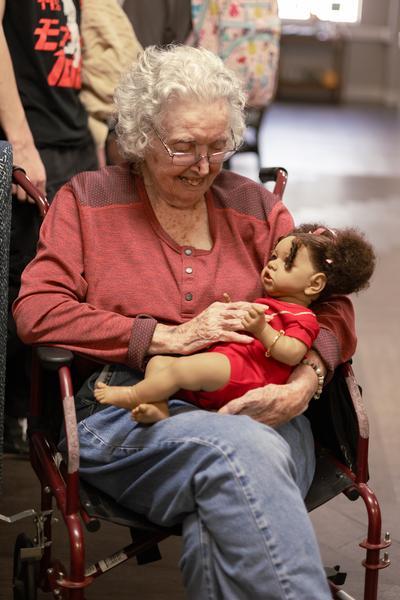 A smiling senior woman in a wheelchair cradling a baby doll in a red outfit