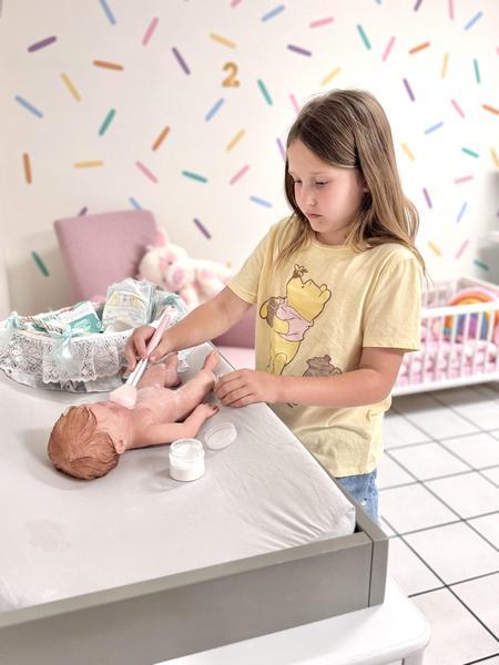 A young girl gently powders a lifelike baby doll on a changing table in a pastel nursery