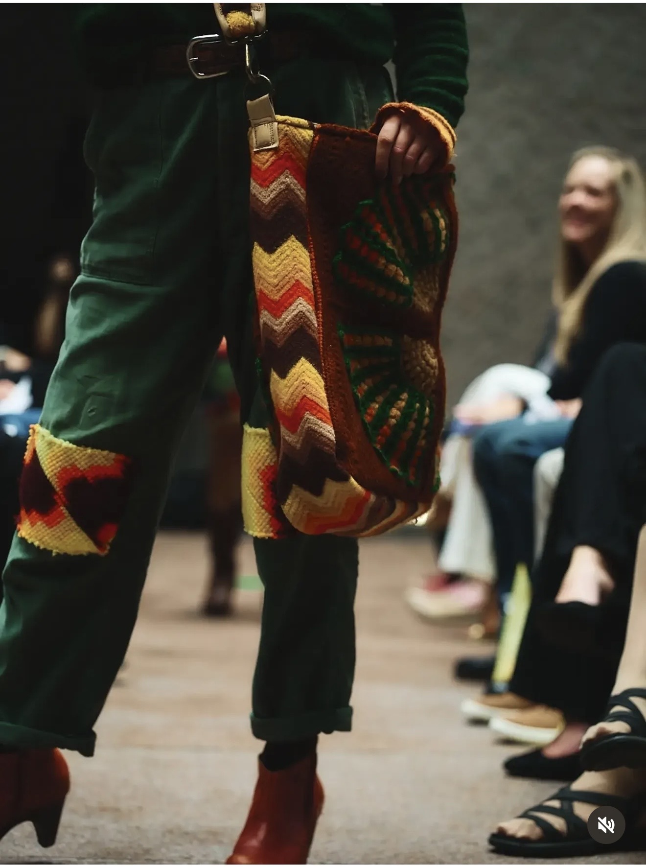 Close-up of a chevron hand-crochet shoulder bag in autumn tones on the runway, worn with patched green trousers and red boots
