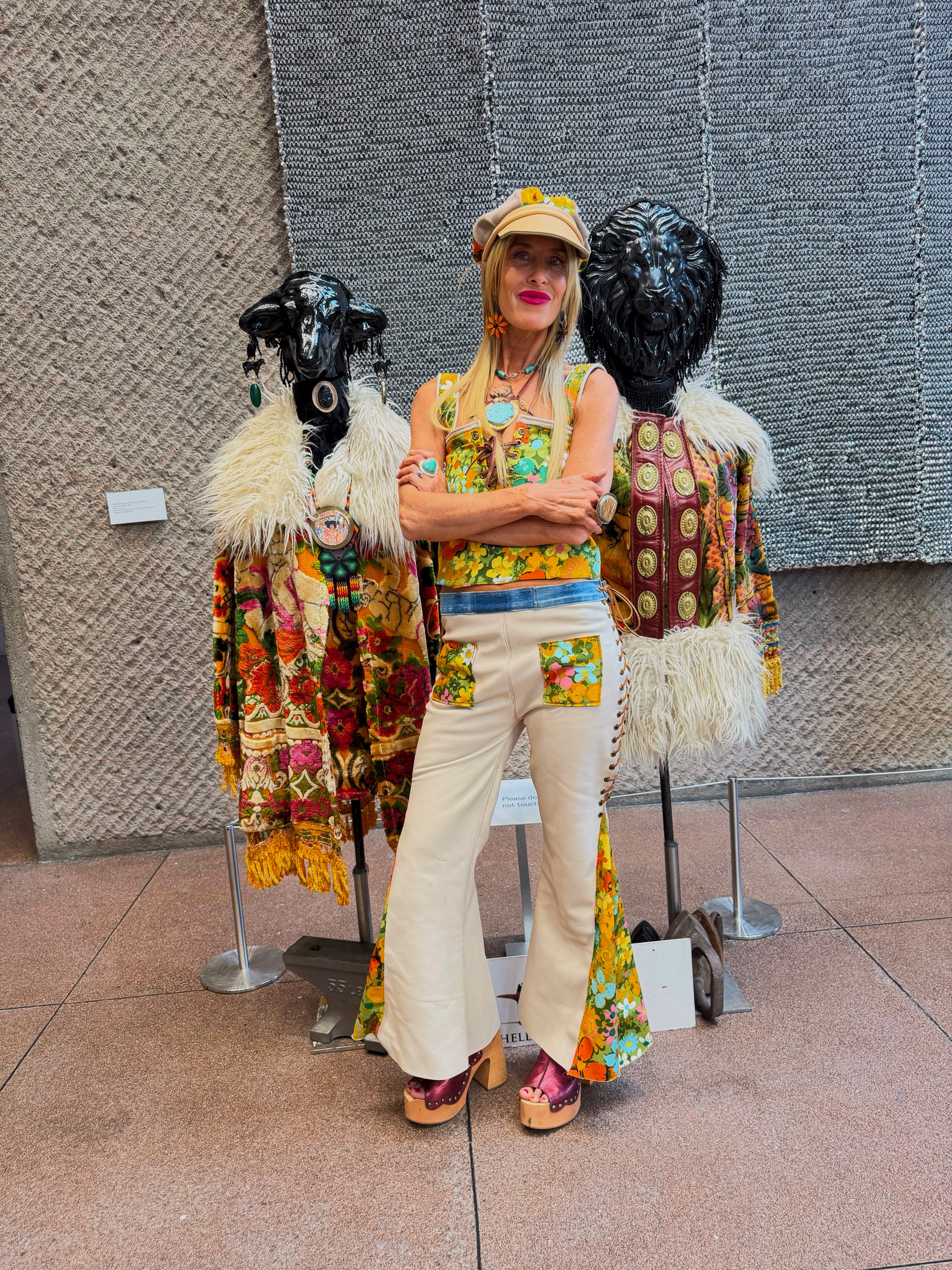 Michelle DaRin in a floral lace-up top and cream flared leather pants with turquoise jewelry, between two black animal-head sculptures at a gallery