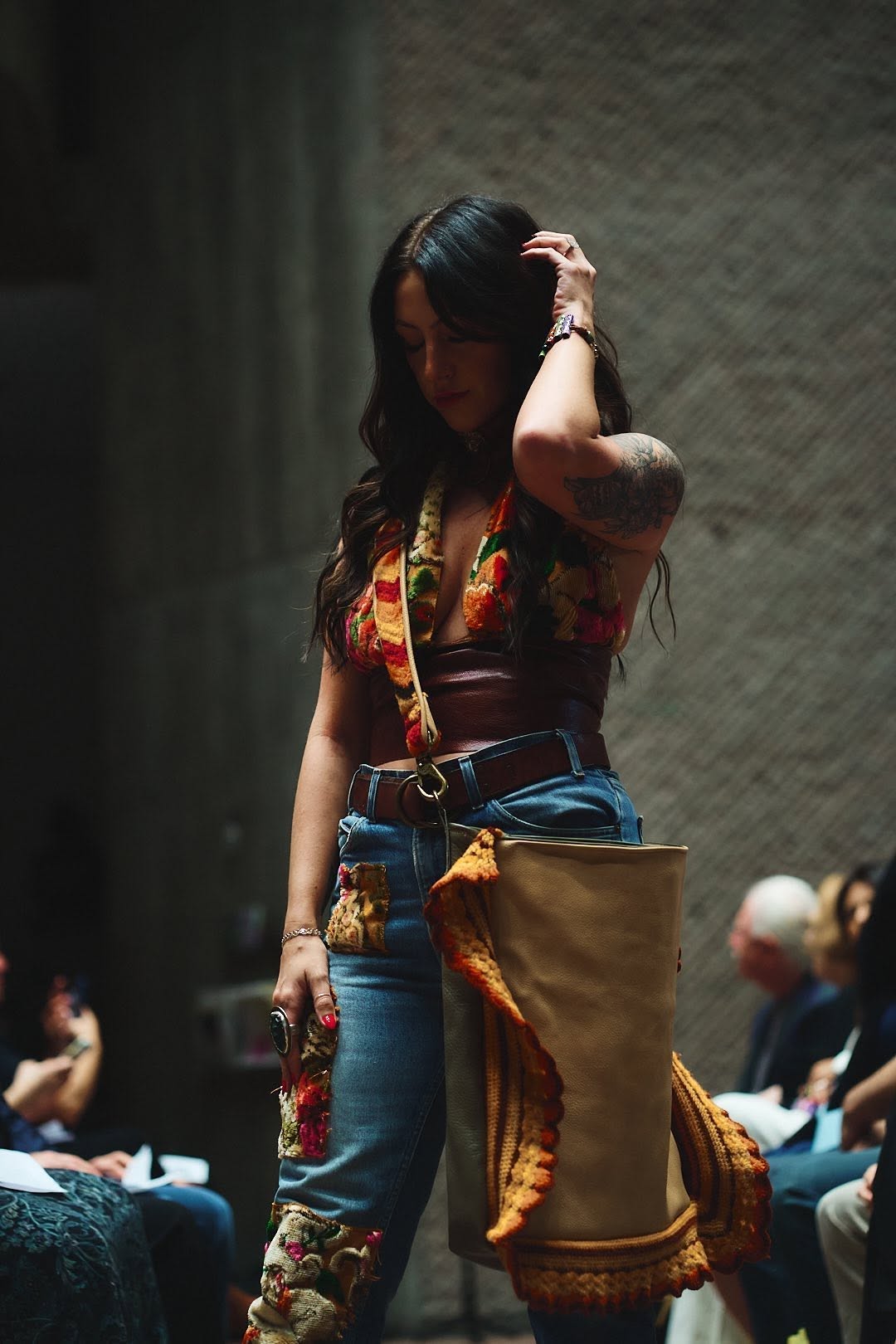 Runway model carrying a tan leather tote with hand-crochet trim, styled with patchwork floral jeans and a leather corset belt