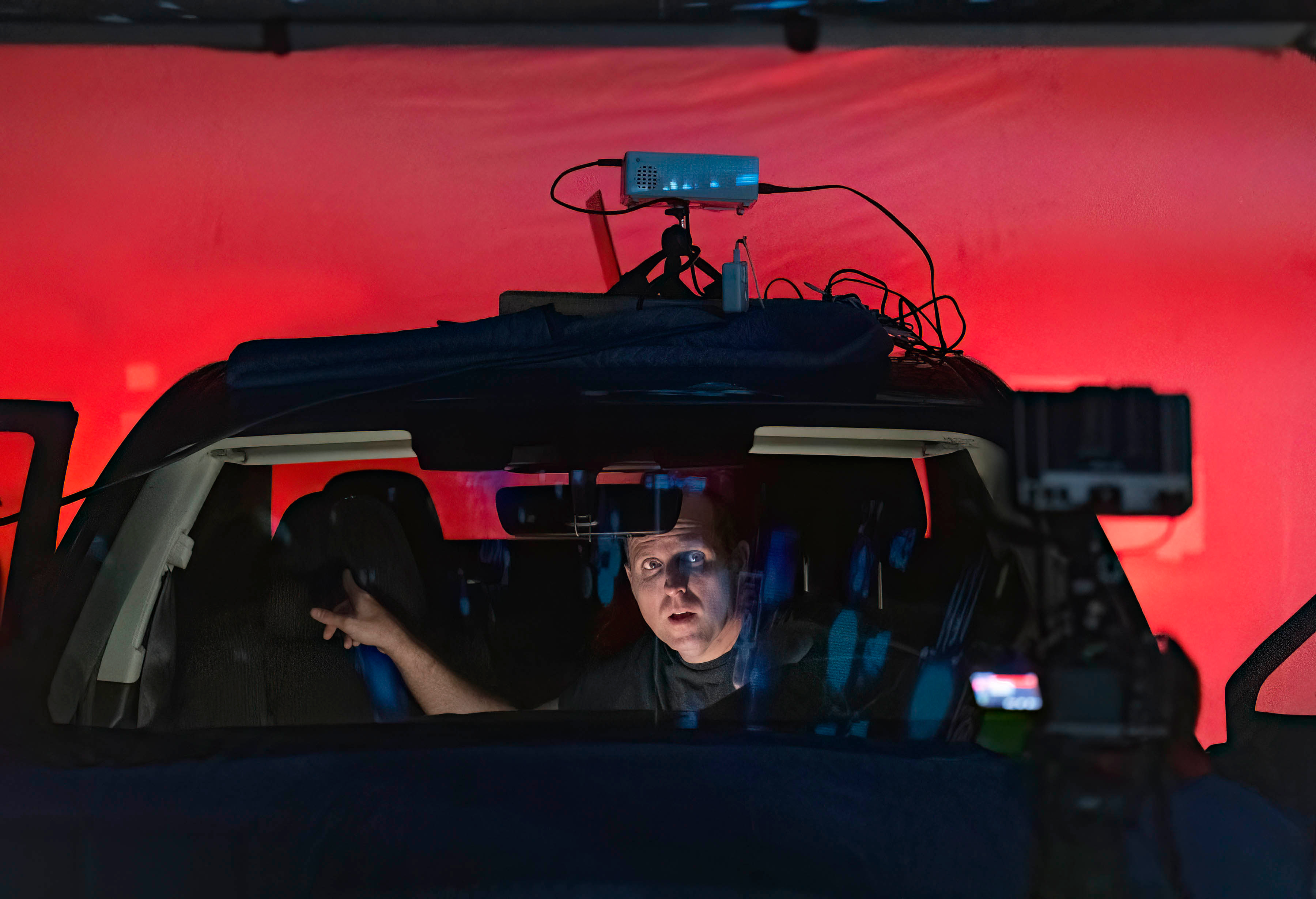 An actor performing in a car rig against a red backdrop with a projector mounted on the roof