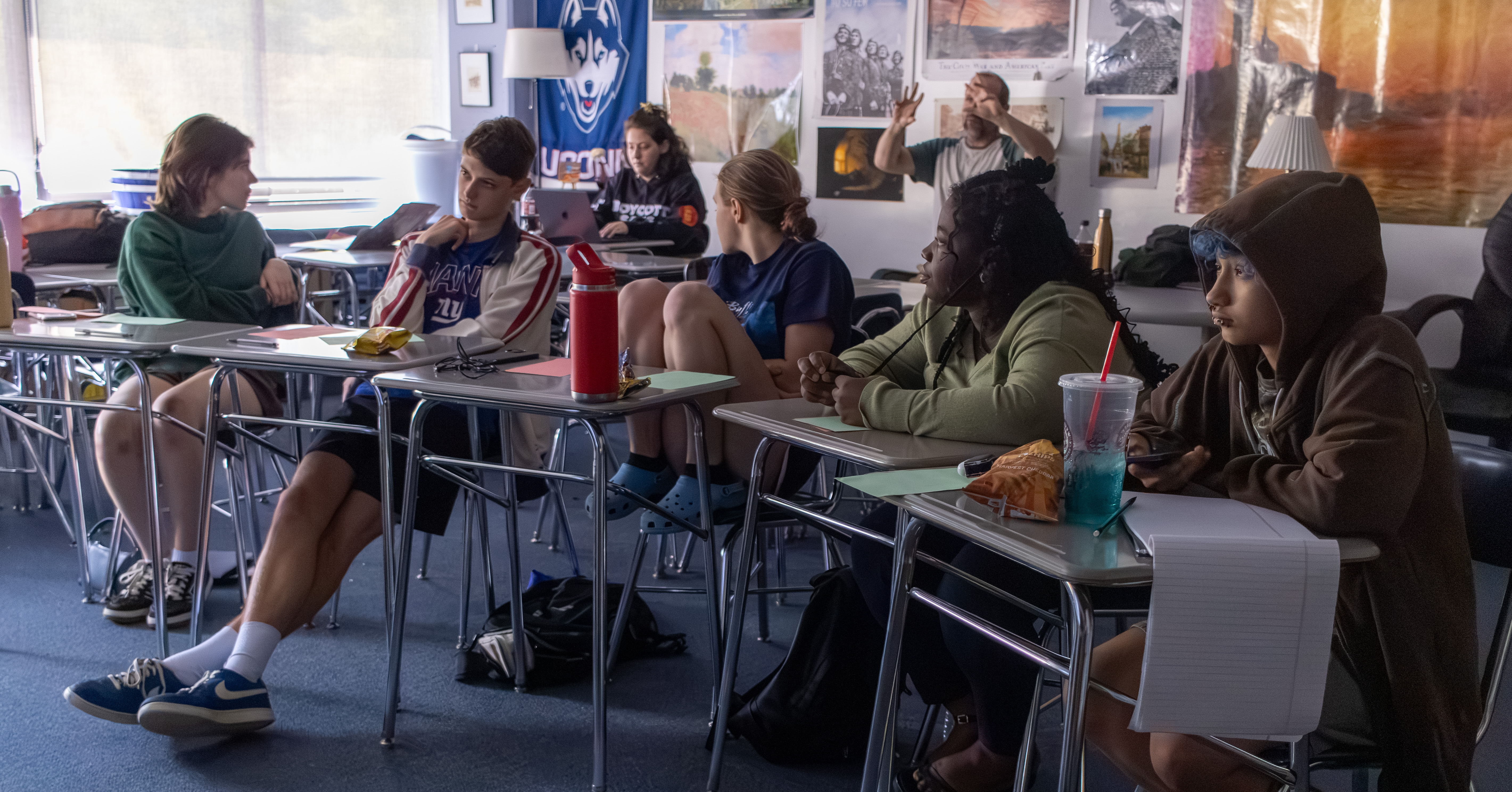 A full room of teen writers seated at desks, mid-discussion with a leader gesturing at the front