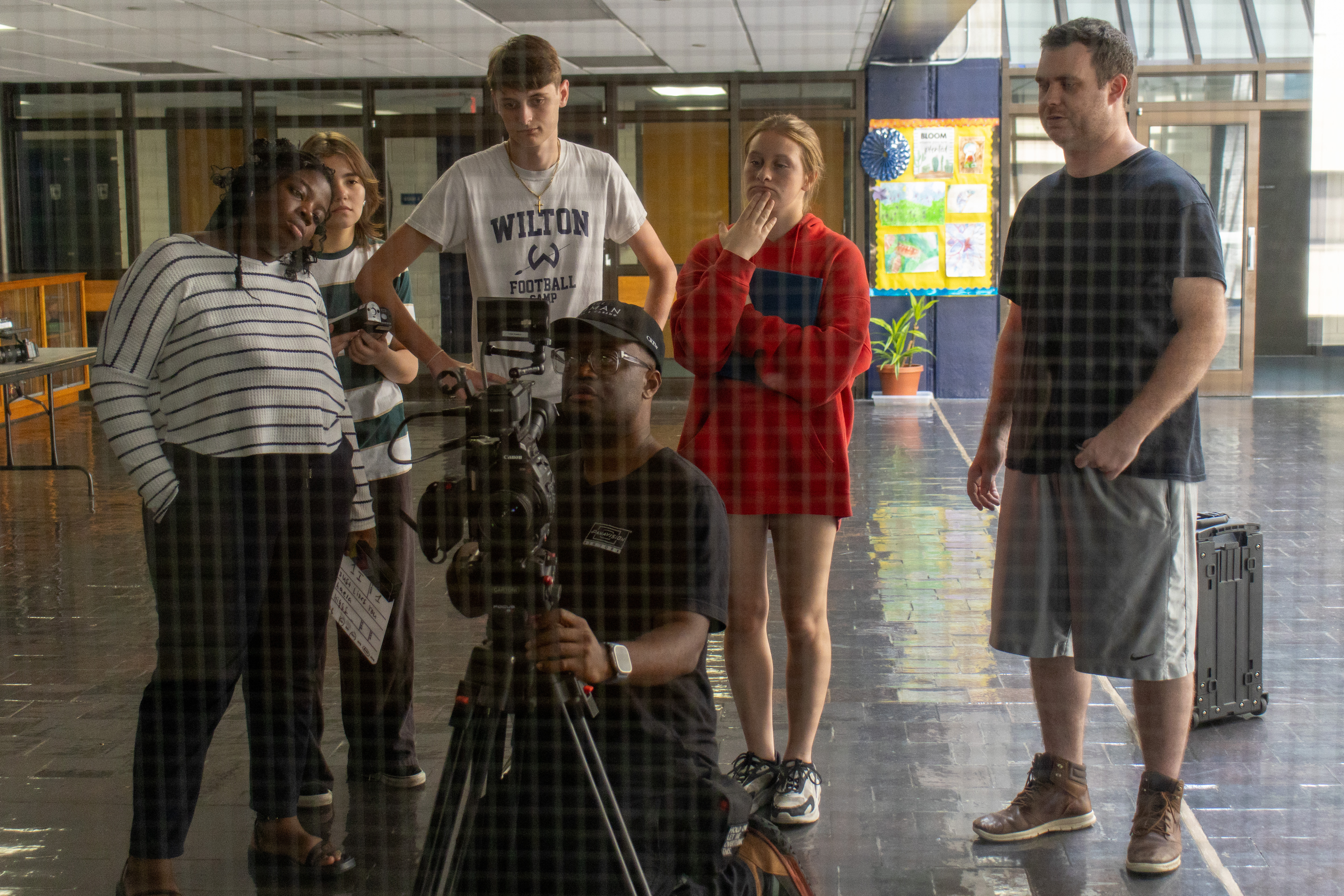 Students and an instructor gather around the camera in a school hallway between setups
