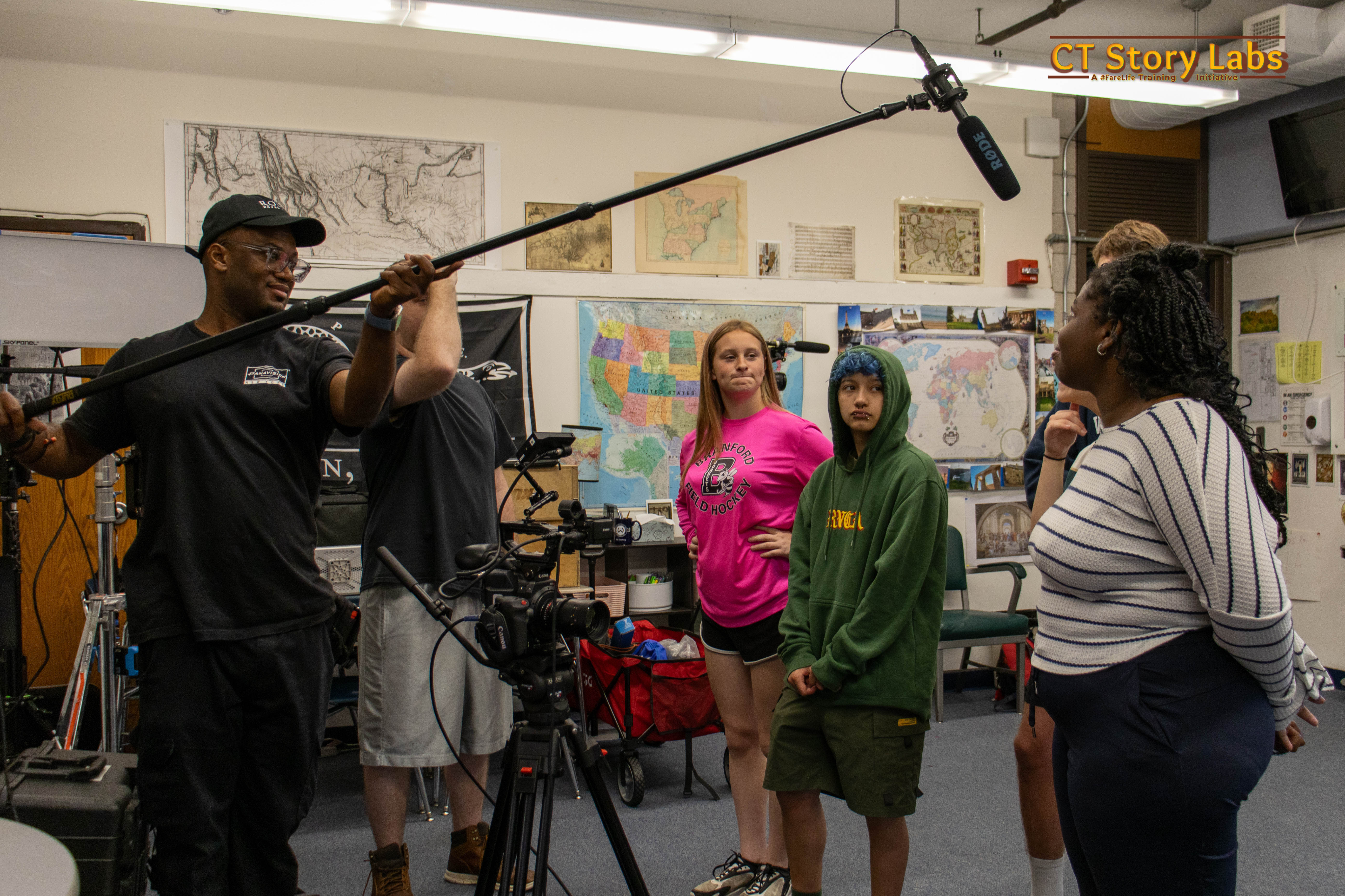 A student operating a boom microphone on set while the crew directs a scene