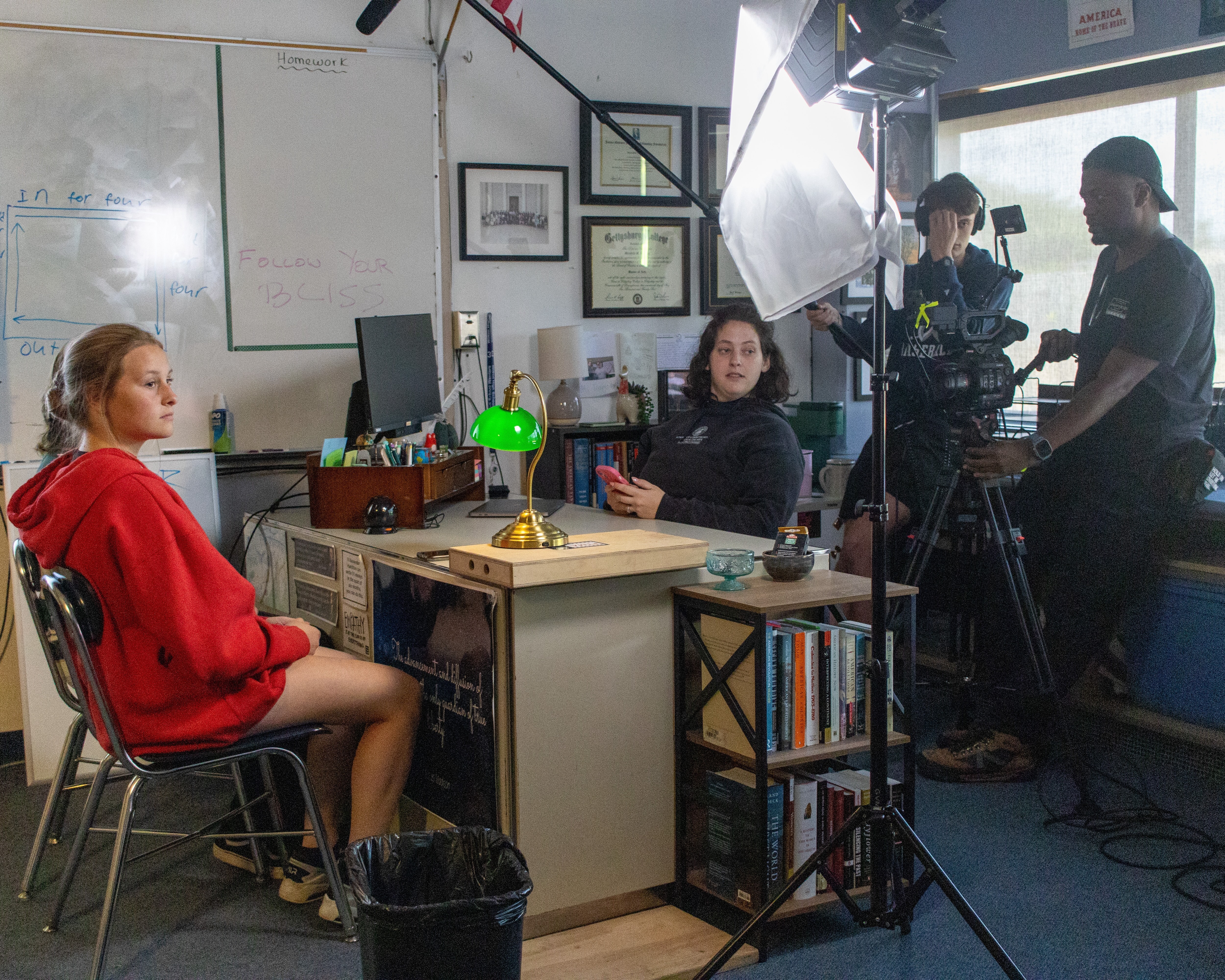 A student crew lights and films an actor for an interview-style scene in a classroom