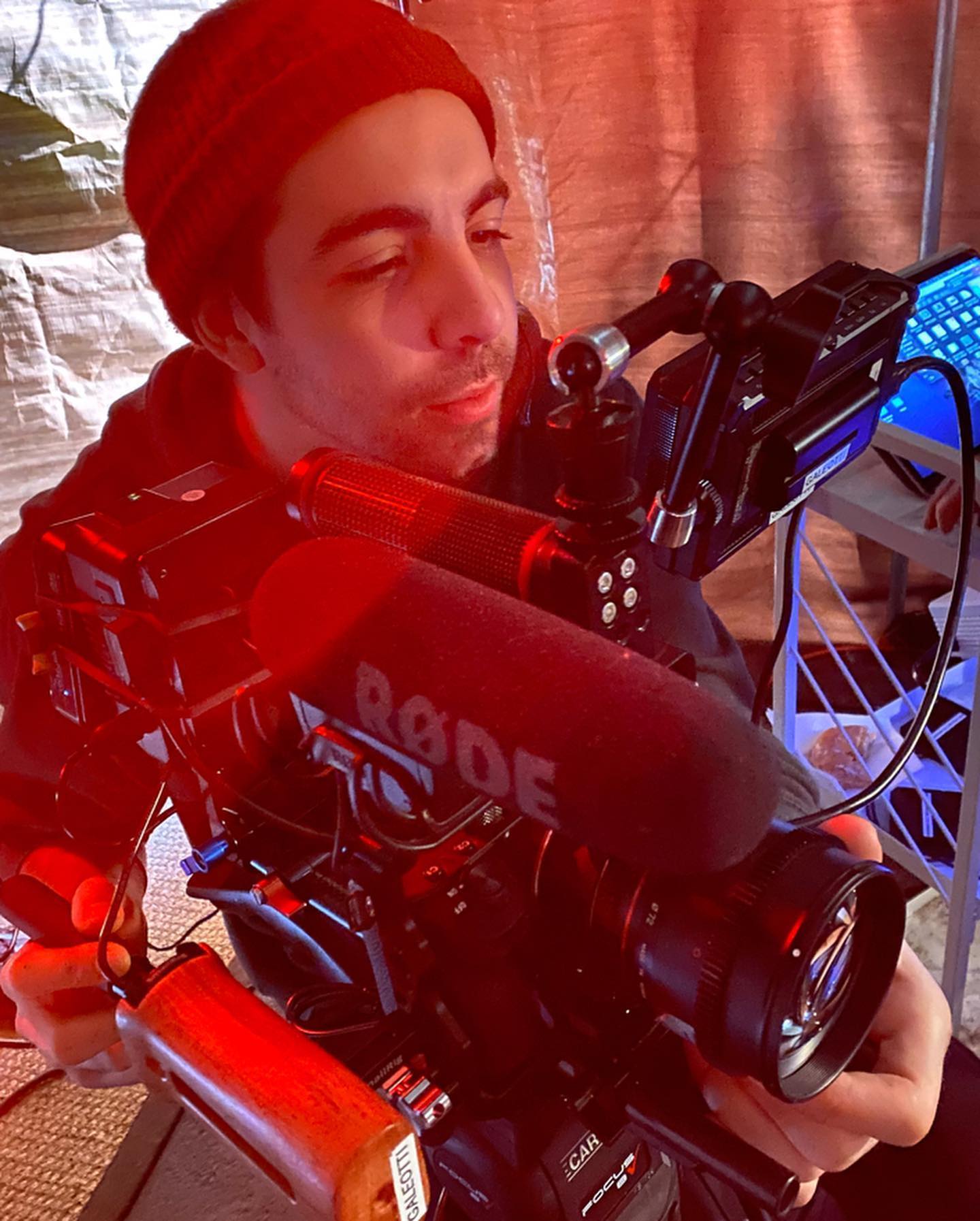 A camera operator in a red beanie framing a shot with a rigged cinema camera and boom mic