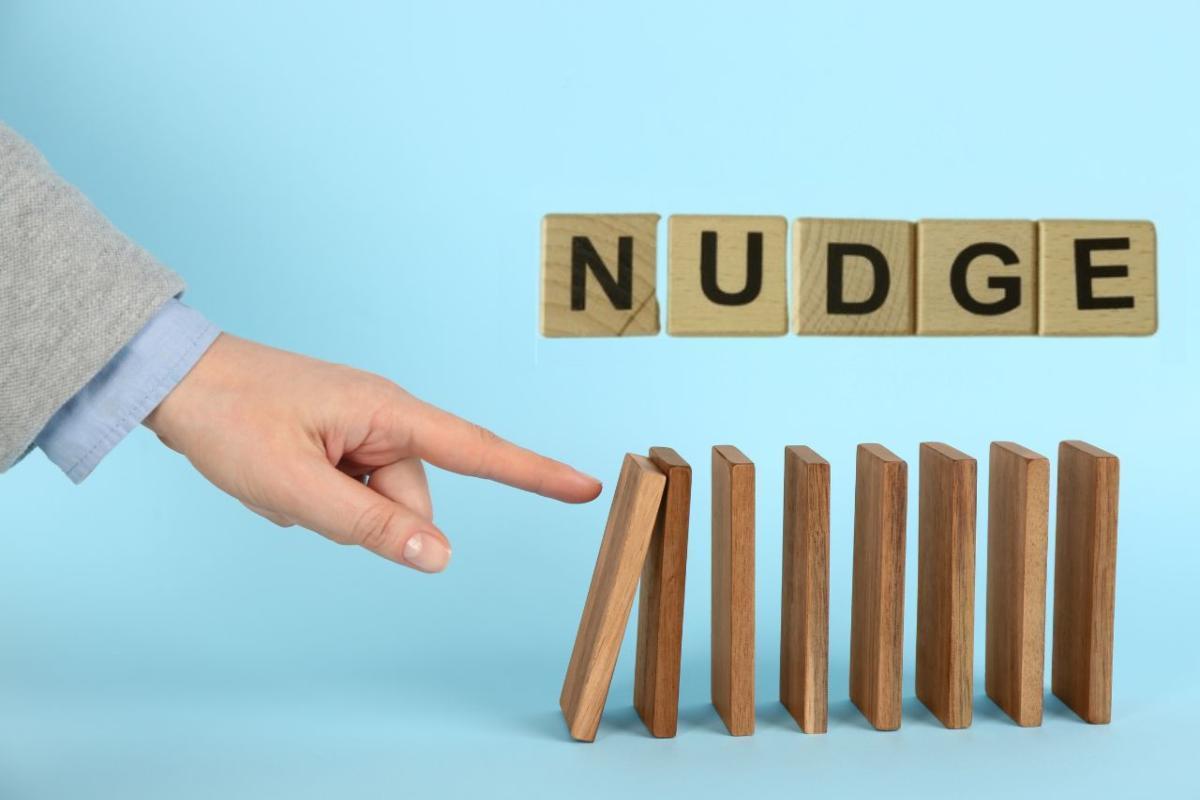 A finger nudging the first in a row of wooden dominoes beneath letter tiles spelling NUDGE
