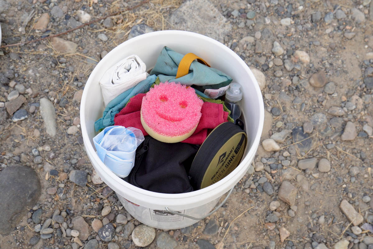 A white 5-gallon bucket on gravel packed with camp kitchen gear including a collapsible sink, sponge, towels and dish soap