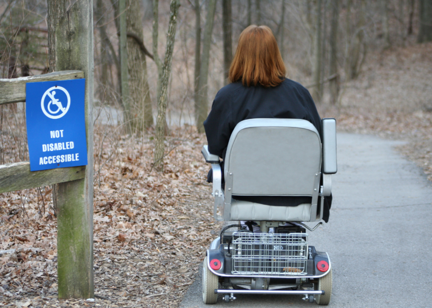 woman in a wheelchair in a forest near a sign which says: not disabled accessible