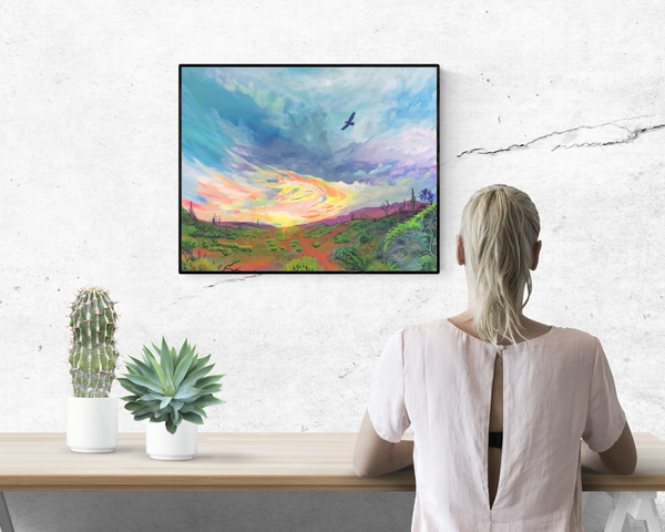 Woman sitting at a table with cactuses and a framed painting on the wall of an Arizona desert sunset.