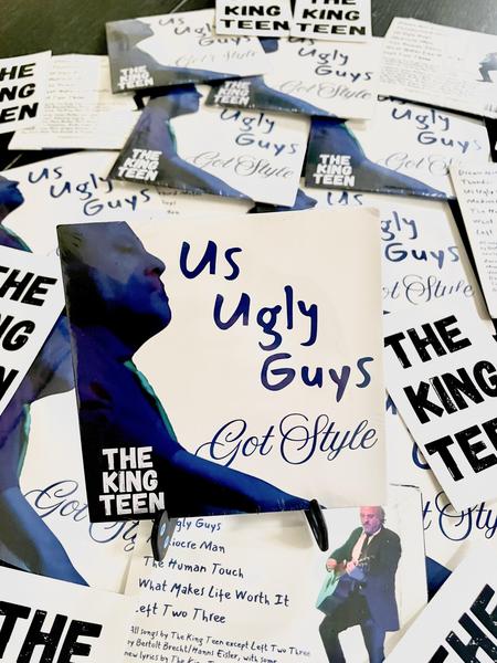 A copy of the CD of Us Ugly Guys Got Style on a little stand, sitting on a bed of other copies of the CD strewn about, plus some stickers that say The King Teen