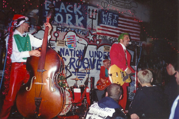 Onstage photo of a band in front of a wall covered in graffiti words and images. The singer has a red suit and green hair. The other players are dressed in red and green and their are xmas decorations dressed on the standup bass, mic stands, drum kit etc. Some young punks are crowding the front of the stage.
