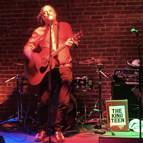 Photo of an incredibly handsome man singing at a microphone and playing guitar in front of a brick wall, bathed in red light, with a sign on a briefcase next to him that says THE KING TEEN