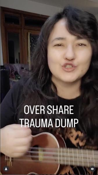 Screenshot from a video showing Erika Kobayashi Libero, who has shoulder length black hair, in closeup, wearing a black T-shirt and playing a ukulele and singing. The words OVER SHARE TRAUMA DUMP appear. 