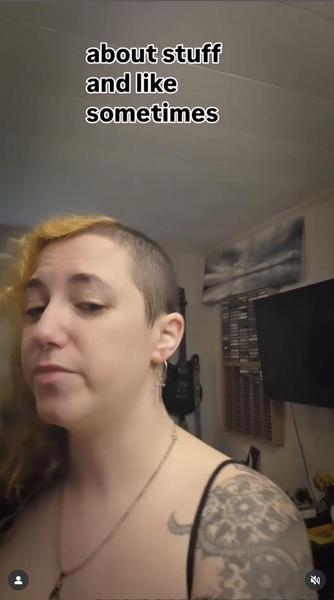 Still from a video showing Em Gee, who has long bleached hair on the right side of their head and a buzzcut on the left, and is wearing a spaghetti strap black top that shows off a tattoo on their left shoulder. Above their head are words from the rolling caption. They say "about stuff and like sometimes."