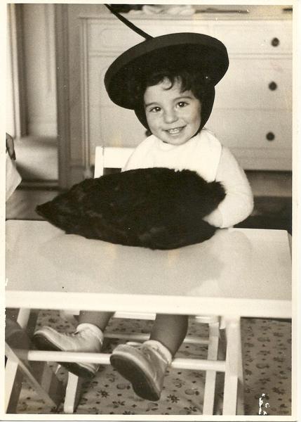 Black and white photo of a toddler boy sitting in a high-chair. He's wearing some kind of round hat with a feather coming out of it and appears to have his hands in a big furry muff or bag of some kind. 