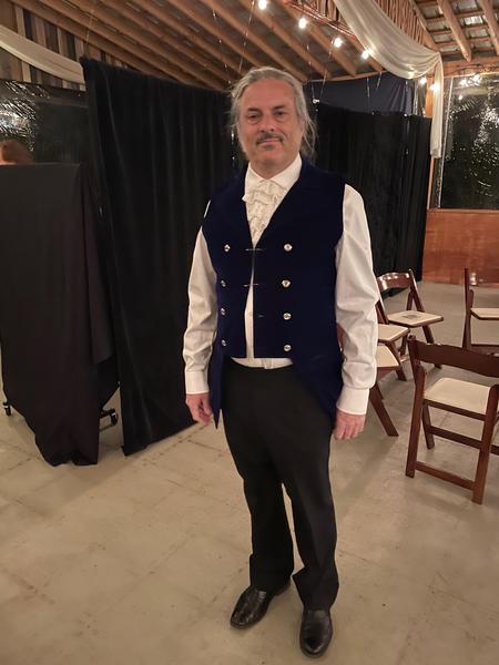 Photo of an incredibly handsome man in a costume of a white shirt, a lacy cravat (I think), a blue vest, black slacks and black shoes. 