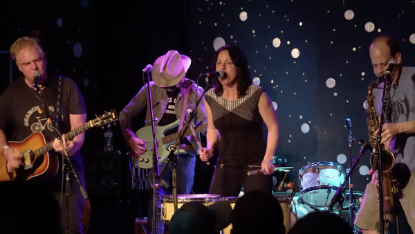 Image of four musicians all playing in a line at the front of a stage. From left, a man singing and playing an electric acoustic guitar, a man playing a bass guitar, a woman standing and playing drums while singing, and a man playing sax. 
