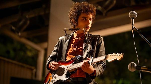 Photo of a guy who looks like Bob Dylan playing an electric guitar and with a harmonica rack on, onstage, wearing a black leather blazer and an orange shirt.