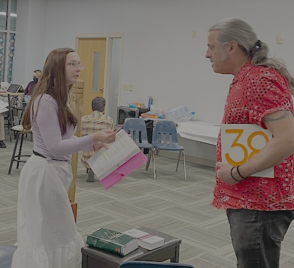 Photo of a woman in a pink shirt and a long white skirt, holding a script in one hand and a business card in the other, talking to a man in a red shirt and black jeans, holding a placard under his arm (it's white with an orange 38 on it, and is standing in for a salver). They are in a large
carpeted room in a school-like setting, with chairs and a few other people in the background. There are some books on a piano bench between the man and woman. 