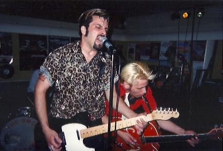 Photo of King wearing a leopard shirt and playing a black Telecaster electric guitar, yelling into a microphone as guitar player Tom Double D Thumb, wearing a red western shirt and white tie and with died blond hair, plays a big Orange Gretsch behind him. Drummer Big Stick Mick is mostly hidden behind King. 