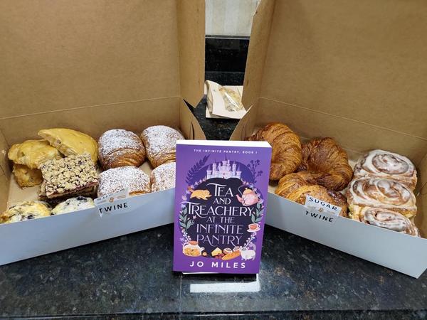 A copy of Tea & Treachery at the Infinite Pantry sitting in front of two boxes filled with croissants, cinnamon buns, scones, and fruit bars