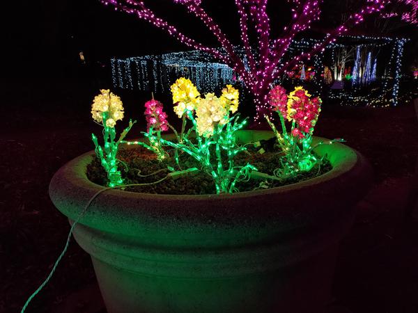 A pot full of lit-up artificial flowers in front of a holiday lights display