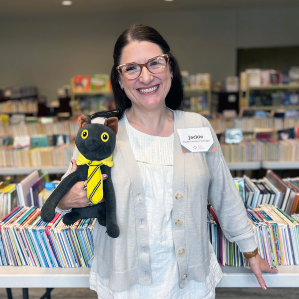 A smiling woman wearing light colored clothes standing in front of a row of books holding a black cat stuffie. The cat has a yellow tie.
